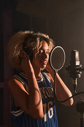 Side view of focused young female singer with Afro hairstyle standing while singing song on mic during rehearsal in professional recording studio - ADSF41727