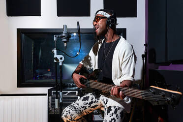 Männlicher afroamerikanischer Musiker mit Dreadlocks spielt E-Gitarre und singt in der Nähe des Mikrofons während einer Probe in einem professionellen Studio - ADSF41758