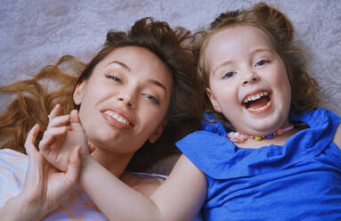 High angle cheerful ute little girl in blue dress lying on bed embracing mother showing care and love looking at camera - ADSF41942