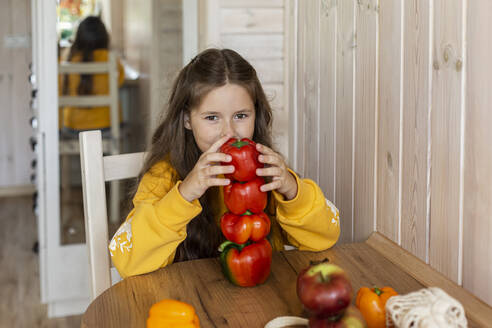 Mädchen sitzt am Küchentisch mit gestapelten roten Paprikaschoten - LLUF00983