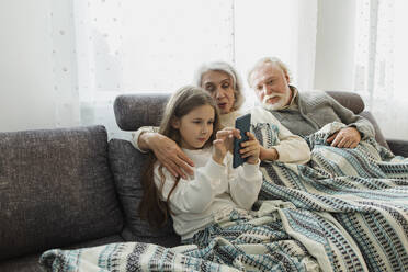 Grandparents and granddaughter sitting on couch under a blanket - LLUF01011