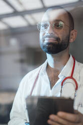 Lächelnder Arzt mit Tablet-PC im Krankenhaus durch Glas gesehen - JOSEF15197
