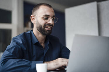 Happy businessman using laptop at desk in office - JOSEF15209