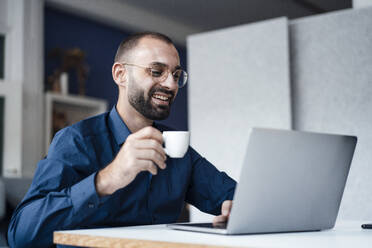 Glücklicher Geschäftsmann mit Kaffeetasse und Laptop am Schreibtisch im Büro - JOSEF15210