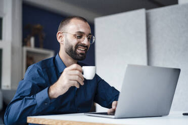 Happy businessman holding coffee cup using laptop at desk in office - JOSEF15210