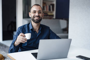 Glücklicher Geschäftsmann mit Kaffeetasse und Laptop am Schreibtisch im Büro - JOSEF15211