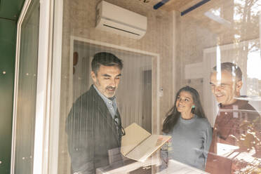 Immobilienmakler bespricht den Kaufvertrag mit den neuen Hausbesitzern, die am Fenster stehen - JCCMF08310