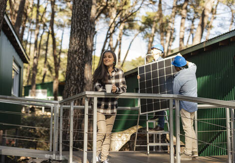 Photovoltaik-Techniker bei der Montage von Solarmodulen auf einem nachhaltigen Haus mit dem Hausbesitzer auf der Veranda - JCCMF08328