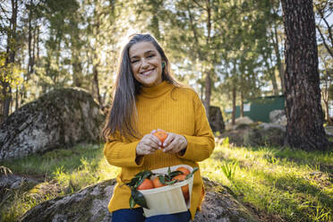 Smiling mature woman holding crate with tangerines - JCCMF08437