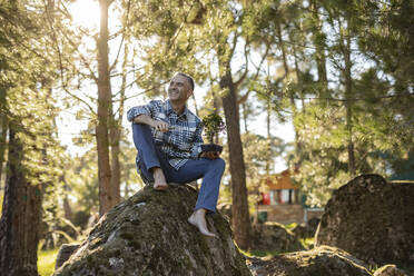 Mature man sitting on a rock holding a bonsai - JCCMF08441