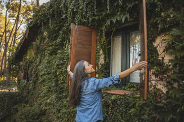Mature woman closing window shutters of house with overgrown facade - JCCMF08496