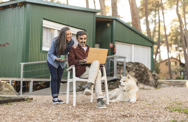 Lächelndes reifes Paar mit Hund und Laptop vor einem grünen Gebäude - JCCMF08503