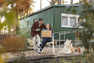 Smiling mature couple with dog and laptop outside green building - JCCMF08504