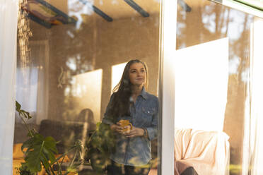 Mature woman holding coffee cup looking out of window of a house - JCCMF08510