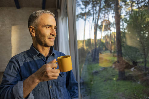 Älterer Mann hält Kaffeetasse zu Hause und schaut aus dem Fenster - JCCMF08521