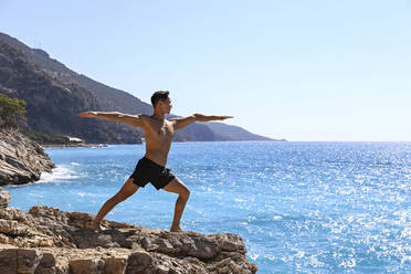 Shirtless Mann übt Virabhadrasana Yoga-Pose stehend auf Felsen in der Nähe von Meer - SYEF00001