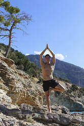 Junger Mann übt auf einem Felsen stehend die Baum-Pose Yoga - SYEF00002