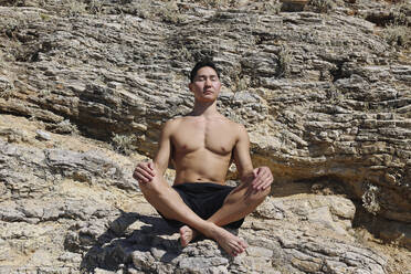 Hemdloser junger Mann mit geschlossenen Augen meditiert auf einem Felsen an einem sonnigen Tag - SYEF00003