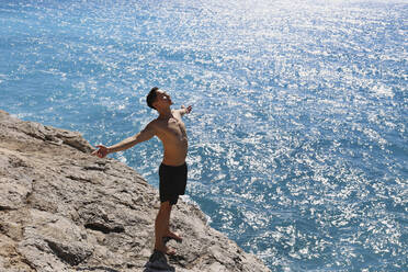 Carefree man with arms outstretched standing on rock by sea - SYEF00004