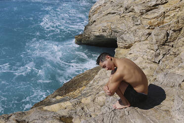 Hemdloser Mann umarmt Knie auf Felsen am Meer - SYEF00006