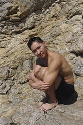 Junger Mann umarmt Knie auf Felsen an einem sonnigen Tag - SYEF00007