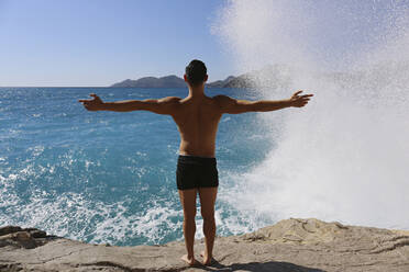 Mann mit ausgestreckten Armen schaut auf Wellen, die auf einen Felsen schlagen - SYEF00008