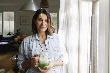 Lächelnde Frau mit einem Glas grünem Smoothie zu Hause stehend - TYF00493