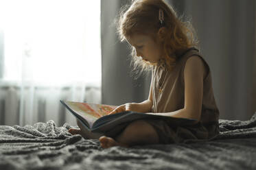 Girl reading book sitting on bed at home - ANAF00610
