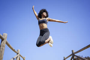 Unbekümmerte junge Frau springt unter blauem Himmel an einem sonnigen Tag - JSMF02585