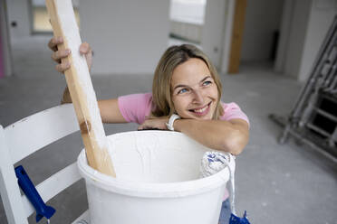 Happy woman day dreaming leaning on paint bucket in apartment - HMEF01493