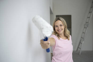 Smiling woman holding paint bucket standing near white wall in apartment - HMEF01494