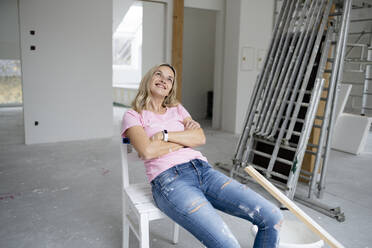 Lächelnde Frau träumt auf einem Stuhl sitzend mit verschränkten Armen in einer Wohnung - HMEF01495