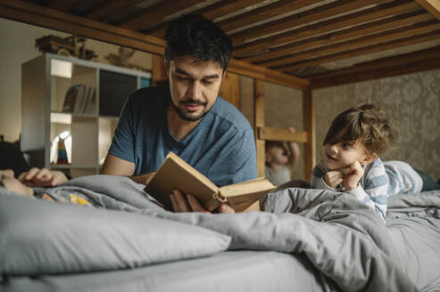 Vater liest seinen Söhnen auf dem Bett zu Hause ein Buch vor - ANAF00625