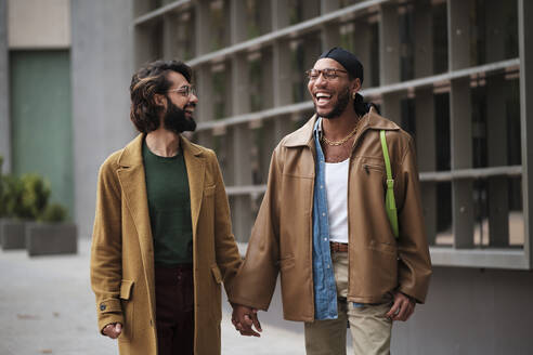 Happy gay couple holding hands walking on footpath - AGOF00305