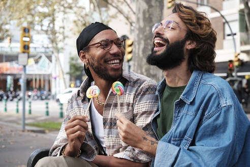 Happy gay couple enjoying candy sitting on bench - AGOF00321