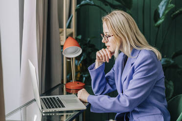 Businesswoman with hand on chin looking at laptop in home office - VSNF00154