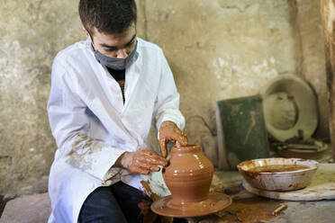 Craftsman wearing protective mask making pot on pottery wheel - JSMF02607