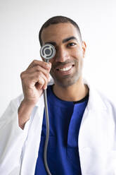 Lächelnder Arzt, der sein Auge mit einem Stethoskop abdeckt, auf weißem Hintergrund - DSIF00632