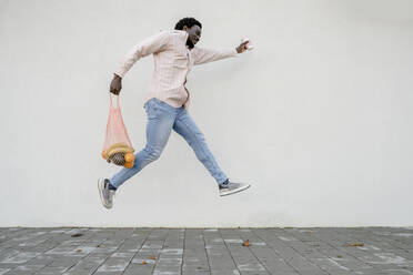 Man with grocery bag and disposable coffee cup jumping in front of white wall - OSF01165