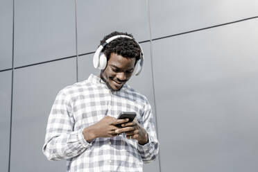 Smiling man wearing headphones using smart phone by wall - OSF01171