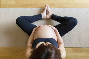 Schwangere Frau mit Händen auf dem Bauch sitzt auf Übungsmatte im Studio - MMPF00574