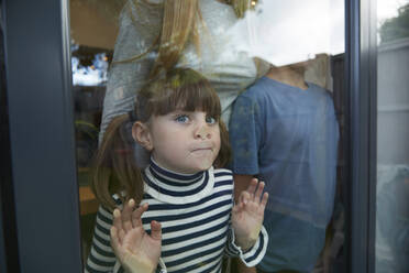 Mädchen, das Nase und Gesicht an ein Glasfenster drückt und mit seiner Familie zu Hause steht - PMF02332