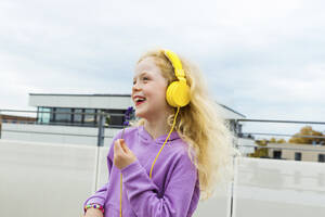 Happy girl holding small flower listening music through headphones at rooftop - AANF00403