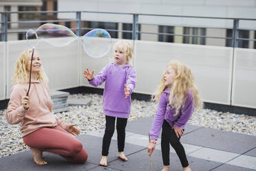 Smiling mother playing with bubble wand by daughters on rooftop - AANF00408