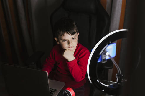 Thoughtful boy sitting with laptop looking at mobile phone in illuminated ring light - LJF02366