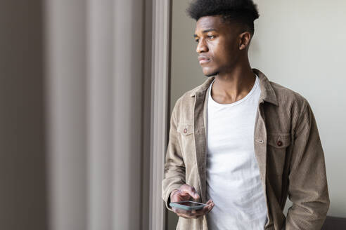 Nachdenklicher junger Mann mit Smartphone, der zu Hause aus dem Fenster schaut - JPTF01134