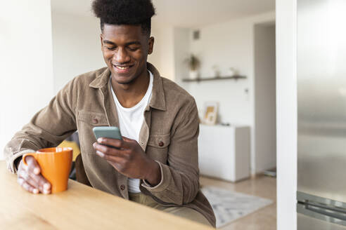 Lächelnder Mann, der sein Smartphone benutzt und einen Kaffee zu Hause trinkt - JPTF01135