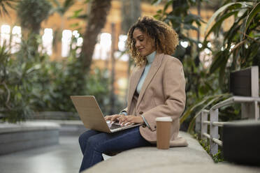 Businesswoman using laptop sitting with disposable cup on bench - JCCMF08531