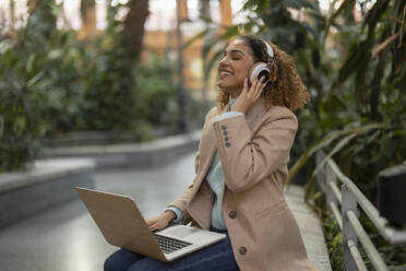 Happy businesswoman listening to music through wireless headphones sitting with laptop on bench - JCCMF08532