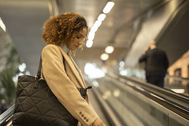 Junge Frau mit lockigem Haar auf Rolltreppe im Bahnhof - JCCMF08545